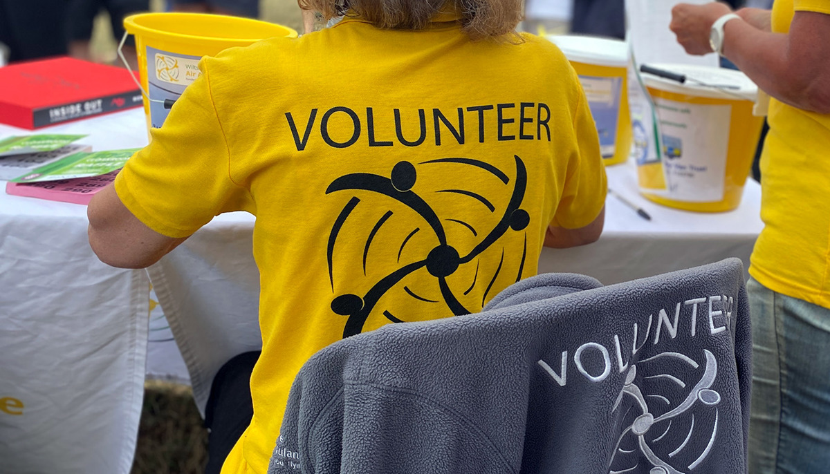 A volunteer at an event wearing a yellow t-shirt, they are sat at a table with yellow collection buckets on and raffle tickets
